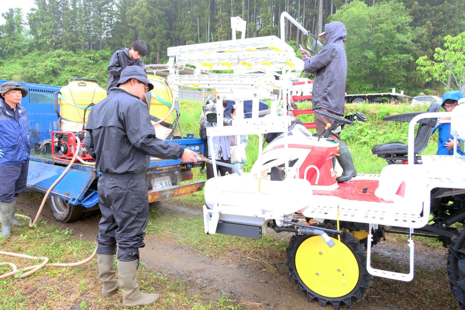 あぜ道に駐車した青いトラックの荷台に積んだ黄色のタンクからのびたホースを持った男性が、田植え機にペースト肥料を供給しており、周りでは数名の人が作業を行っている様子の写真