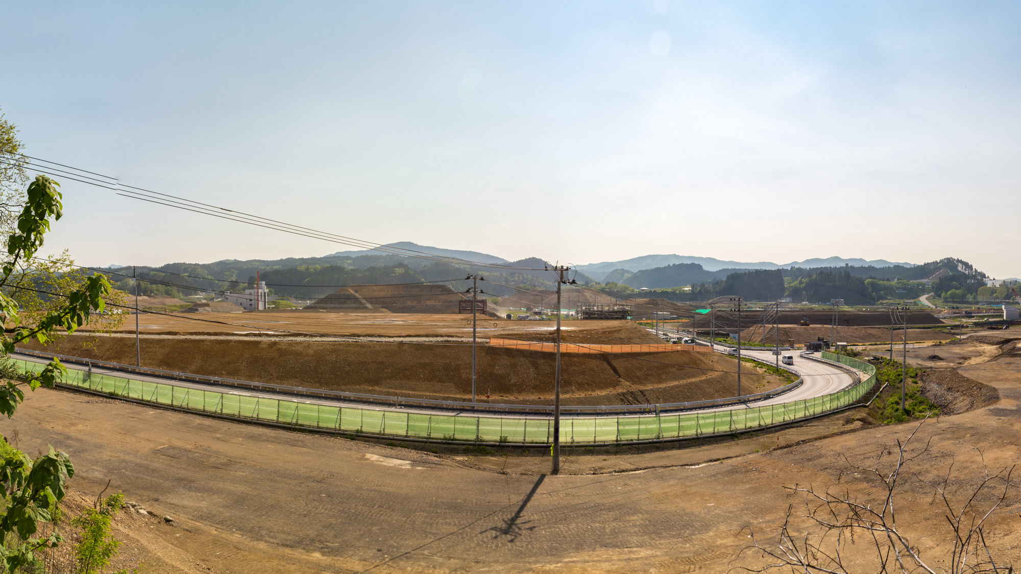 広大な土地を見下ろす定点カメラによる昼間の風景で造成工事が進行中の赤茶けた土の台地で中央には、くねるように伸びた舗装された道路があり、緑色の防音壁のような柵で囲まれている様子を写した写真
