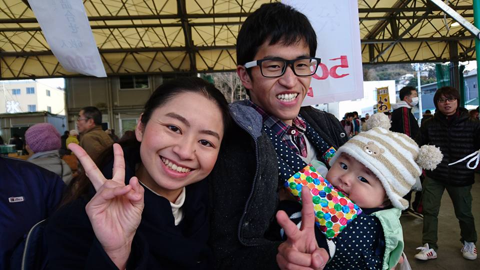 屋根のあるイベント会場で男性が赤ちゃんを抱き、その隣で女性が笑顔でピースサインをしている家族の写真