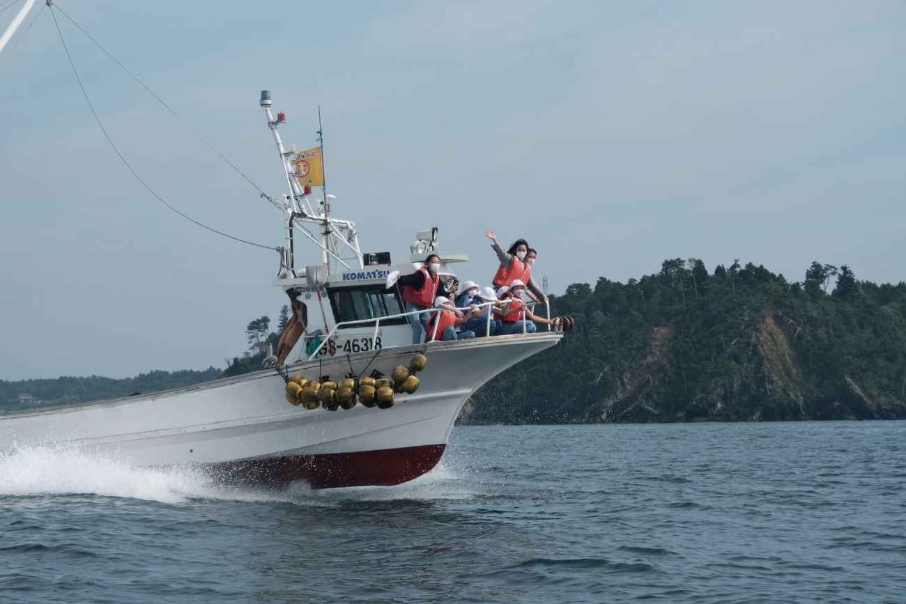 赤い救命胴衣を着た女子学生たちが漁船の先端に乗り、手を振りながら海上での見学を楽しんでいる様子の写真