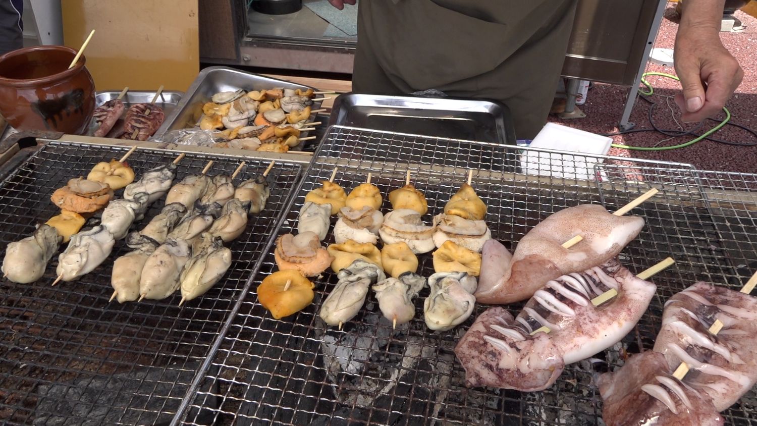 屋外の屋台で、牡蠣やホタテ、ウニなどの串焼きが金網の上で炭火に焼かれており、その横にはイカの姿焼きも並んでいる海鮮炭火焼きの写真