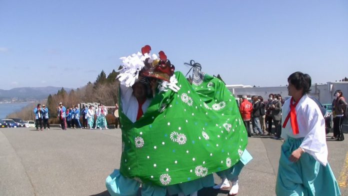 青空の下、獅子舞が道で演舞をしており、鮮やかな緑の布に覆われた獅子が舞う中、白装束の人々が周囲で見守っている様子の写真