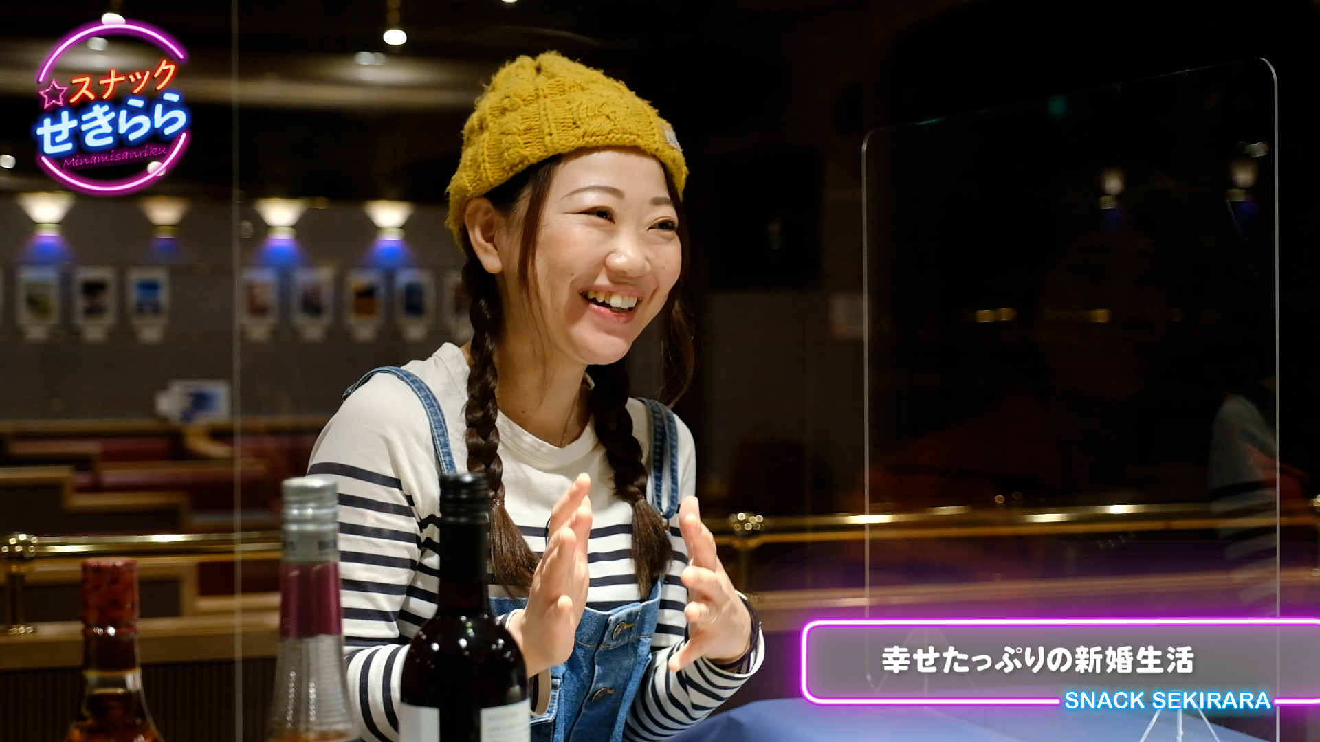 黄色いニット帽をかぶり、ツインテールの髪型で、白地に黒いボーダーの長袖シャツとデニムのオーバーオールを着た女性がカウンター越しに笑顔で話す様子の写真