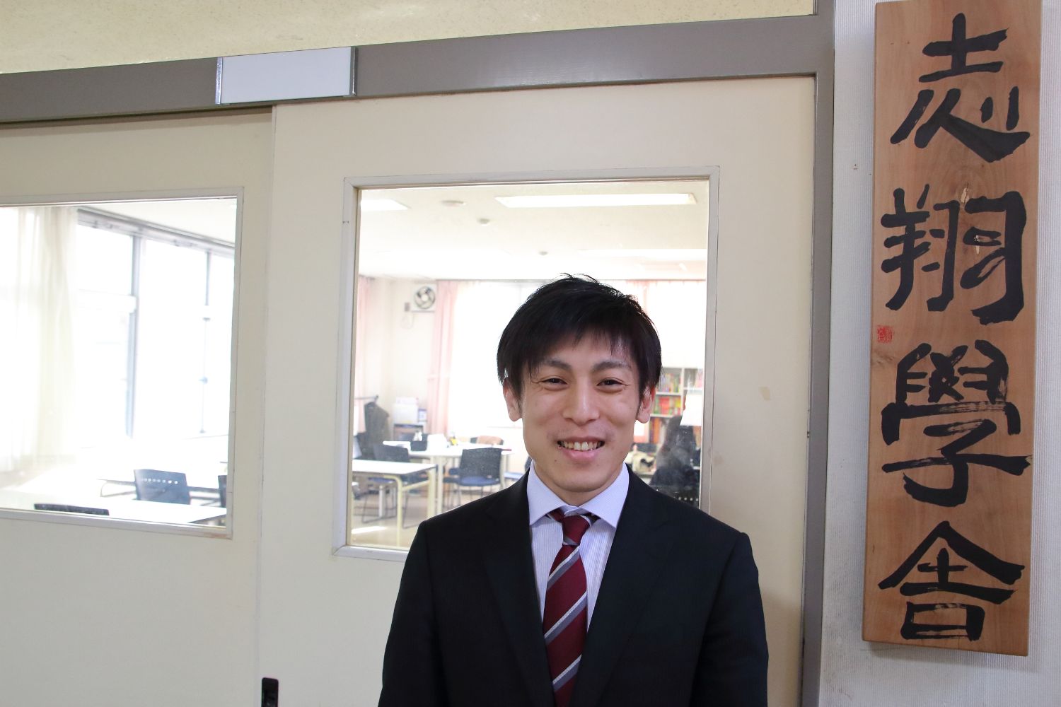 右側には「志翔学舎」と書かれた木製の看板が掲げられた教室の前で、スーツ姿の若い男性が笑顔で引き戸の前に立っている写真