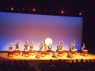 舞台上で、揃いの衣装を着た演者たちが大小さまざまな和太鼓を使って力強く演奏し、手前には観客の姿も見える大森創作太鼓旭ヶ浦（あさひがうら）の郷土芸能発表の写真