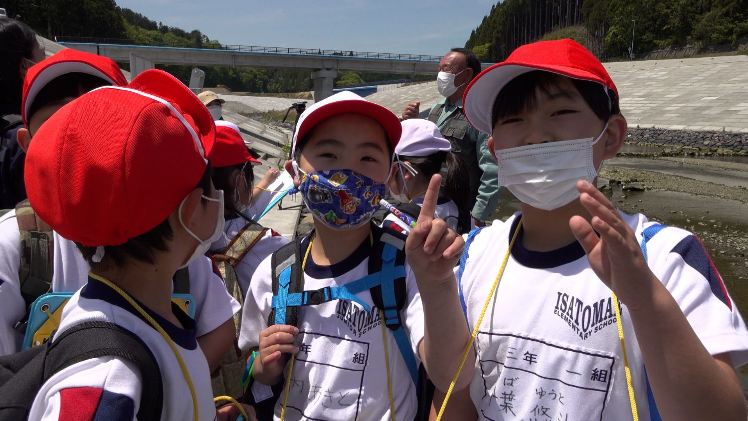 赤白帽子をかぶった小学生の男の子たちが河川敷でマスクを着けて並び、一人の子がカメラに向かって人差し指をあげたポーズをしている写真