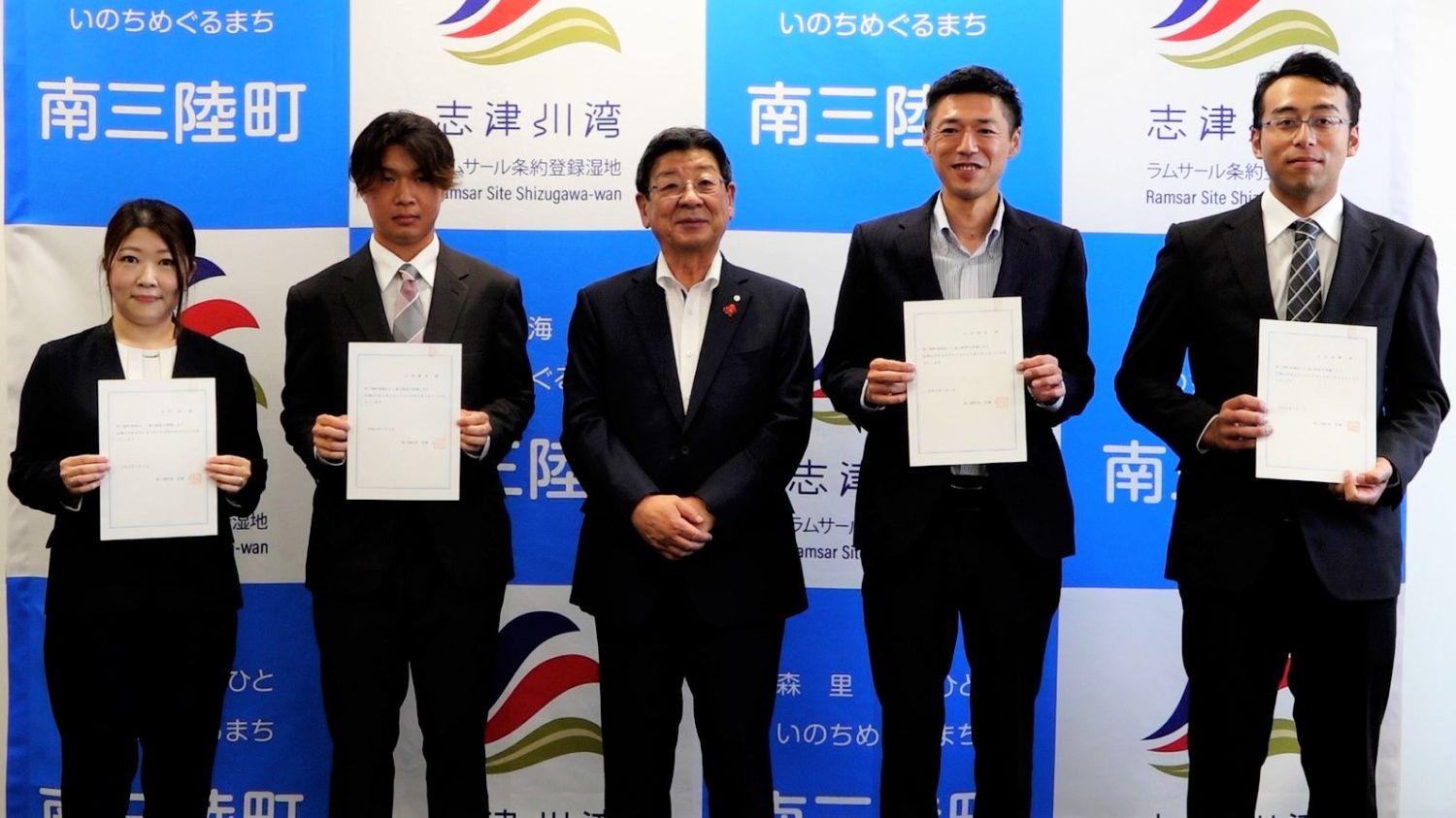 南三陸町のパネル前で地域おこし協力隊の委嘱状を持った太田一さんと協力隊のみなさんが並んでいる写真