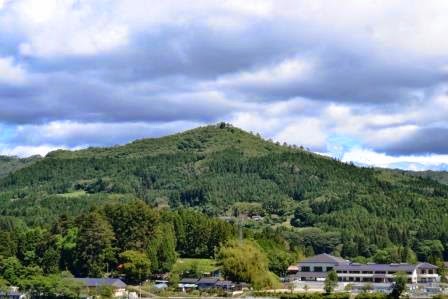 画面中央にあるこんもりとした緑の山を背景にして、手前に学校のような白い建物と森が広がっている山里の写真