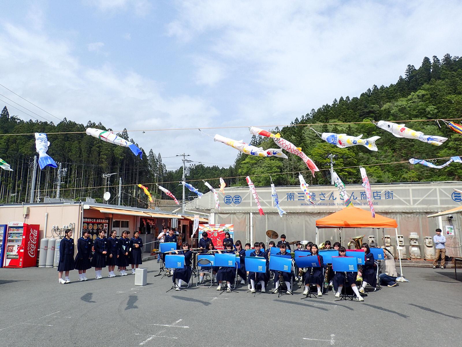 南三陸さんさん商店街の前で、頭上にたくさんのこいのぼりが並ぶ中、吹奏楽部の生徒たちが楽器を演奏し、左側に一列に並んだ女子生徒たちが合唱をしている写真