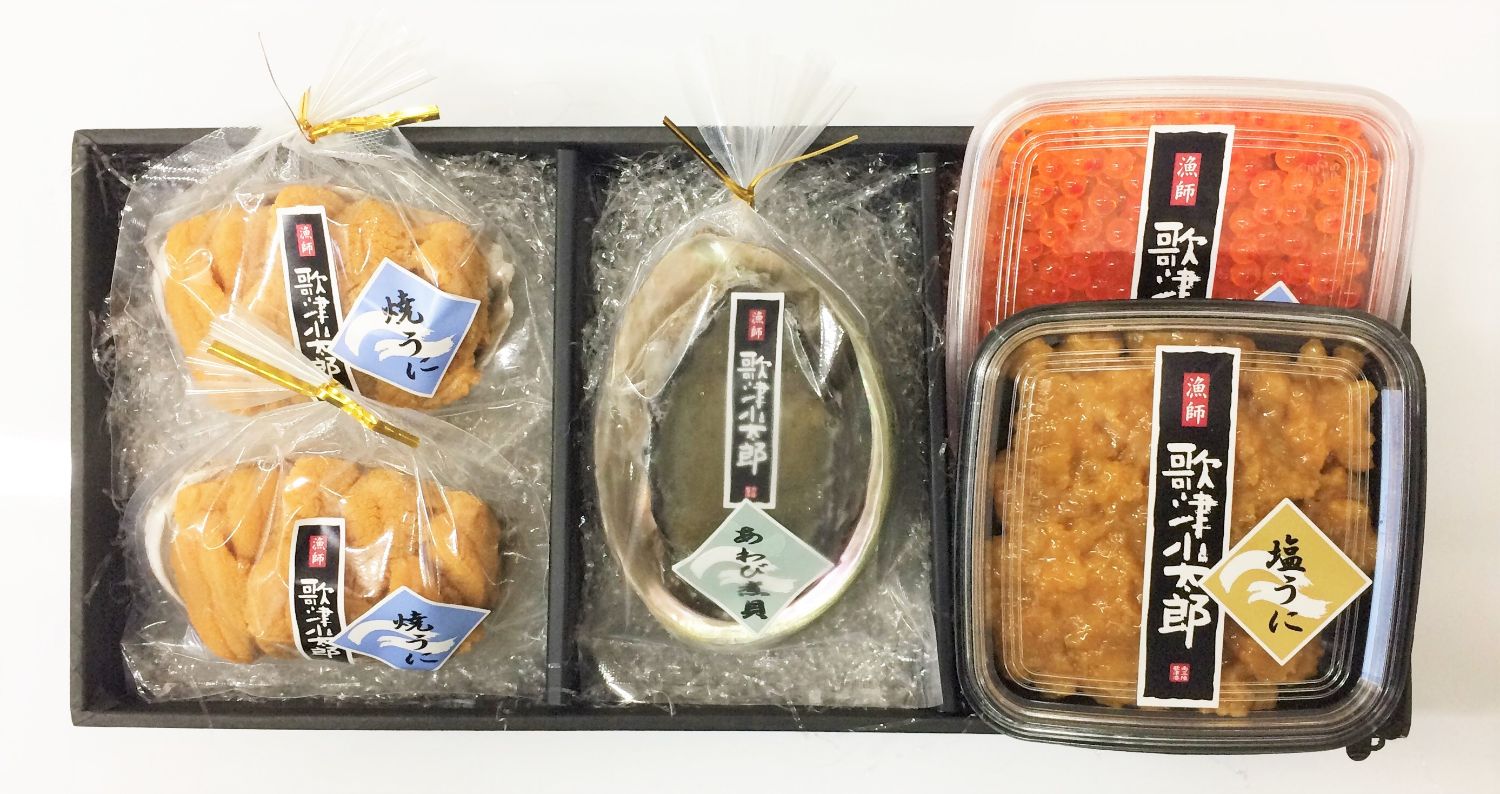 透明の袋に入った焼きウニやアワビ、そしてパックに詰められたイクラと塩ウニが黒い化粧箱に詰め合わされている、高級海産物のギフトセットの写真