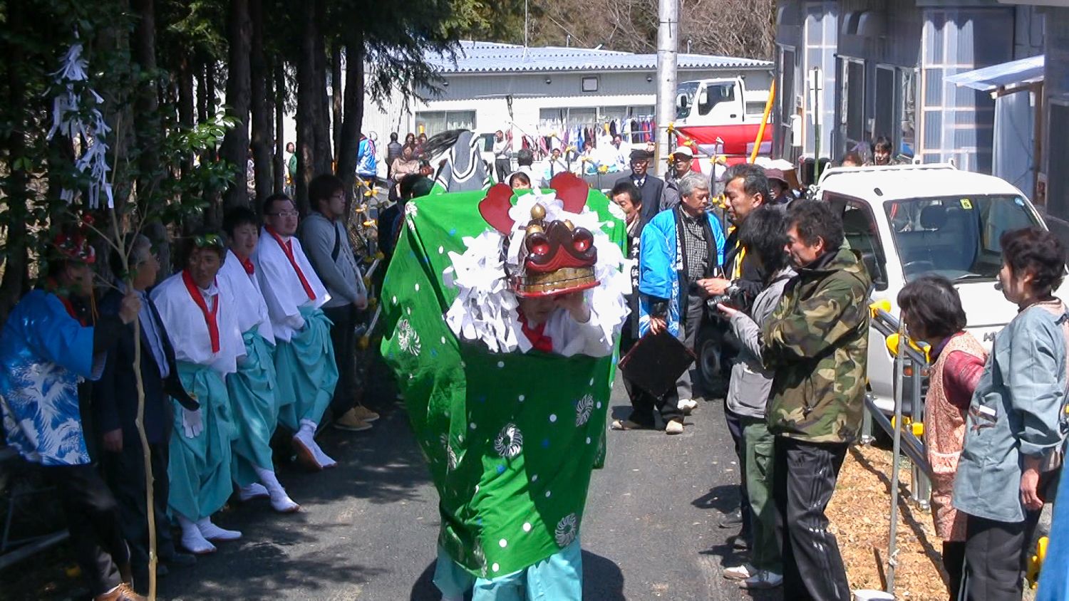 木立の中の細い道で、白装束に水色の袴を履いた人々が並ぶ中、緑の布に覆われた獅子舞が中央を進んでいる様子の写真