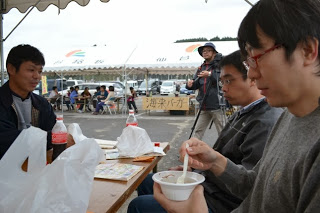 野外のテントの下で、数名の男性が白い容器の料理を手に黙々と食べている様子の写真