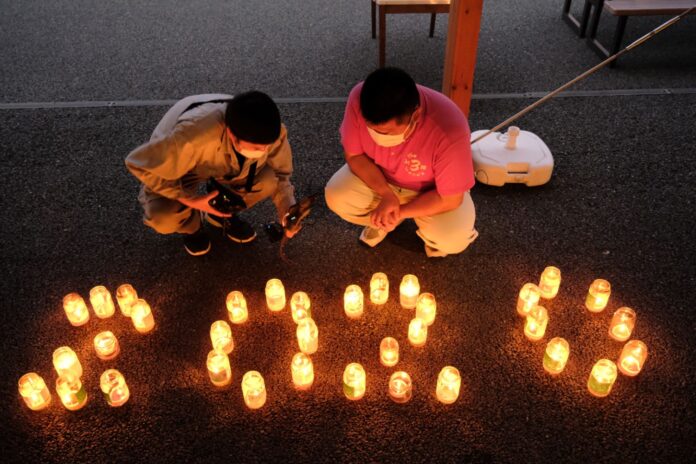 暗くなった地面に置かれたたくさんのガラス瓶のキャンドルが「2020」と形作られており、そのそばで2人の男性がしゃがんで撮影している夜の風景の写真