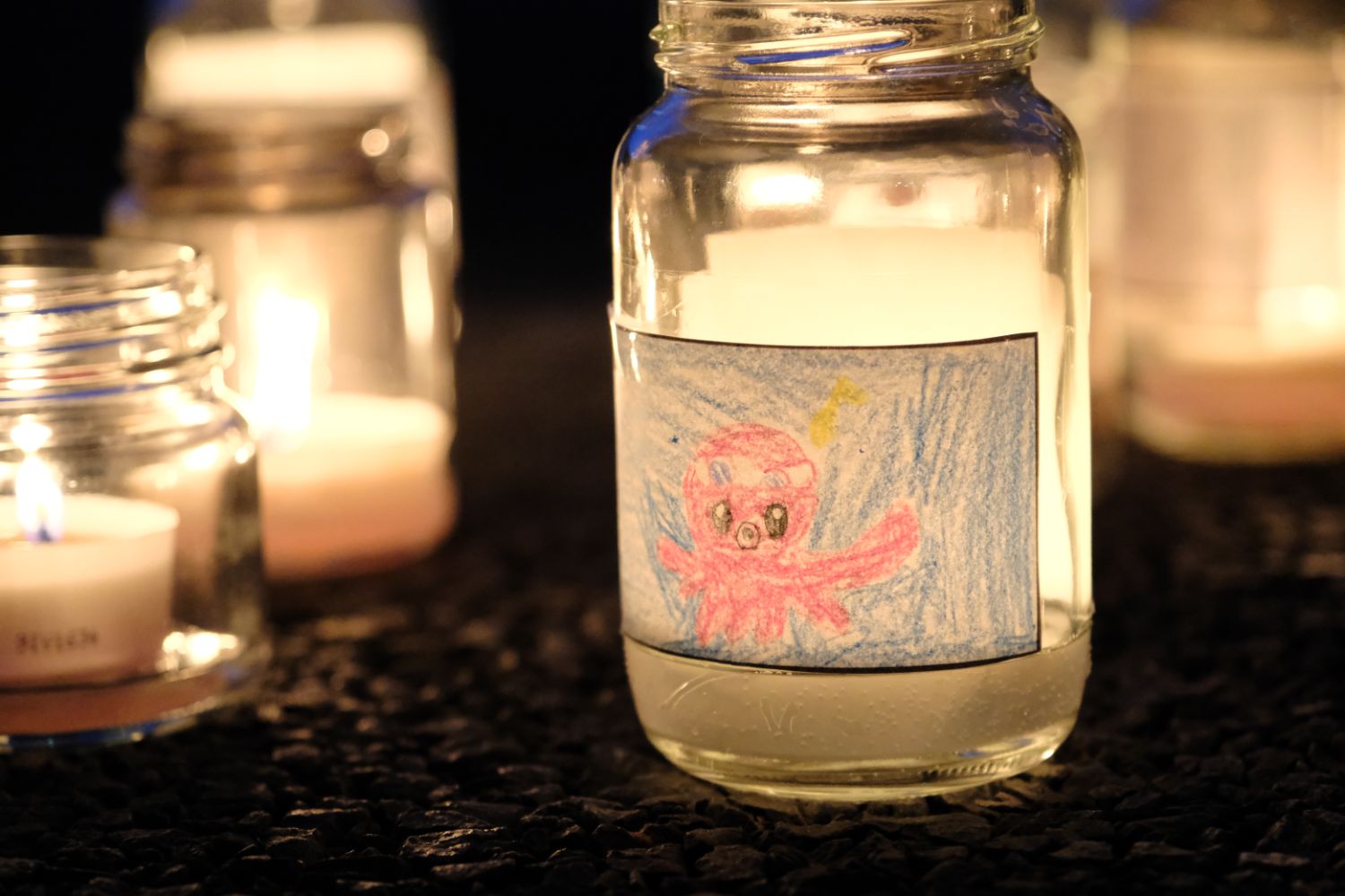 小学生が描いたタコの絵が貼られたガラス瓶の中にキャンドルが灯っており、夜の暗がりに温かな光を放っている様子の写真