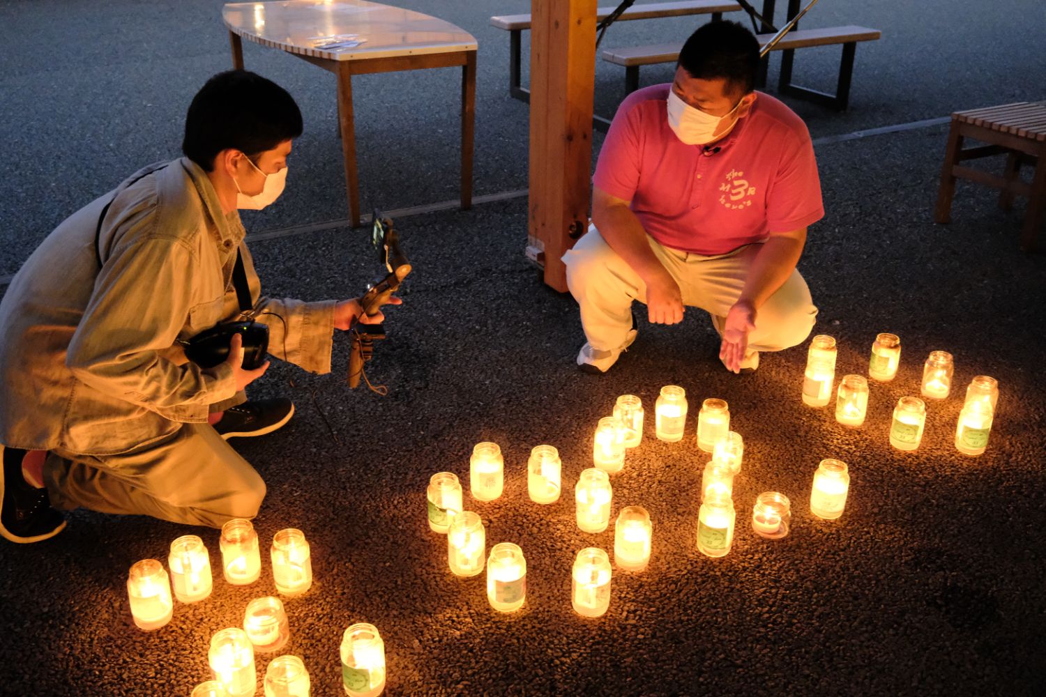 マスクをした2人の男性が、手描きの絵が貼られたガラス瓶のキャンドルで形作られ「2020」」の文字を撮影しながら紹介している写真