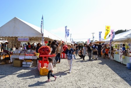 白いテントが左右に並ぶ屋外の会場で、オクトパス君の飾りが置かれたブースの前を子どもと女性が歩き、周囲には多くの人々が買い物や飲食を楽しんでいる祭り会場の様子の写真