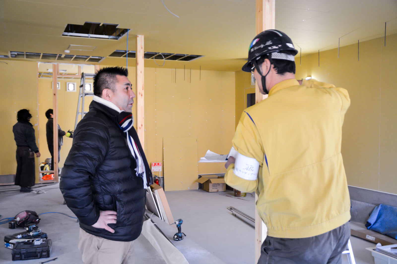 建築中の室内で黒いジャンパー姿の後藤さんが黄色い作業着を着て黒いヘルメットをかぶった男性と話している様子の写真