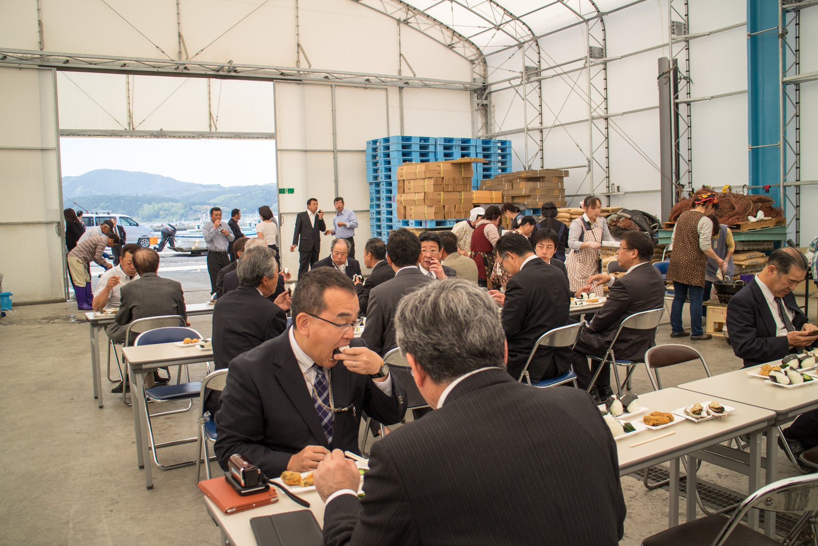 天井の高い大きなテントの中で、スーツ姿の多くの男性がテーブルについておにぎりやカキフライを食べながら談笑し、奥ではエプロン姿の女性たちが配膳や準備をしている写真