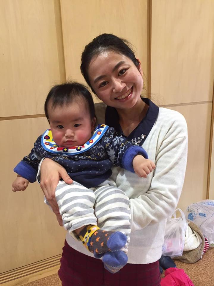 屋内で若い女性が縞模様のズボンを履いた赤ちゃんを両腕で抱っこして笑顔を見せている写真