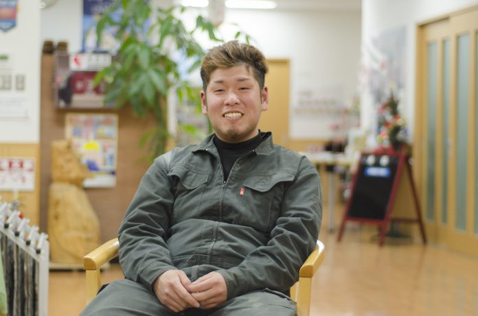 作業服を着た千葉さんが室内の椅子に座り、笑顔でカメラを見ている写真