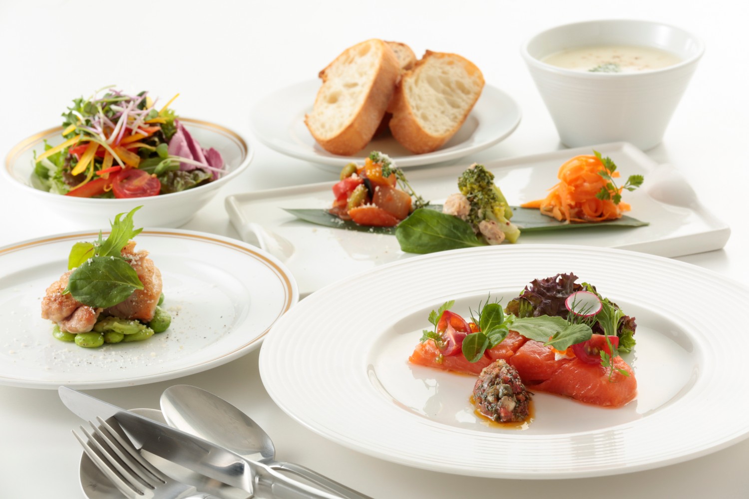 サラダや前菜、銀鮭のコンフィやスープなどが白いテーブル上に盛り付けられた「わたすの贅沢ランチコース」の料理が美しく並べられた、彩り豊かな洋風ランチ料理の写真