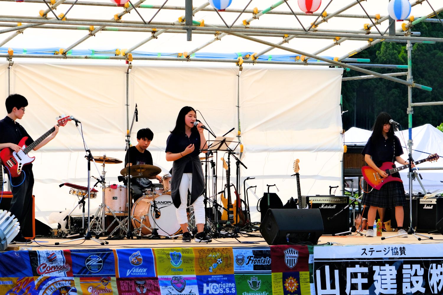 屋外の特設ステージで若者のバンドが演奏し、女性ボーカルが歌っている地域の夏祭りの音楽イベントの写真