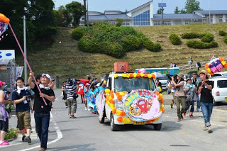色とりどりの飾りや看板をつけた軽ワゴン車が、郵便ポストを載せて沿道をパレードしている、返還式の様子の写真
