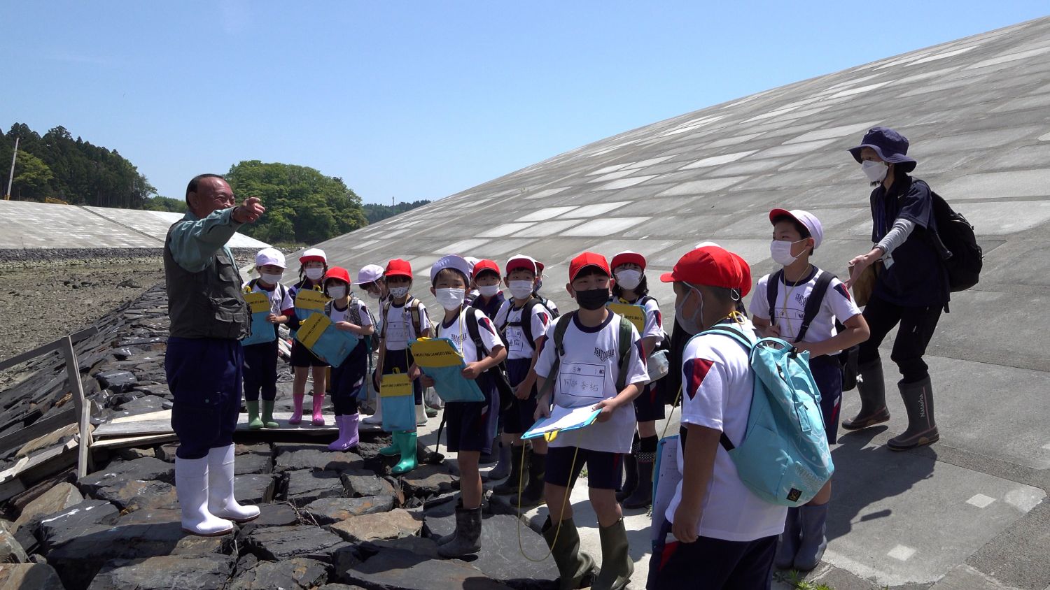 コンクリート造りの堤防の斜面で、年配の男性が指を差して説明し、それを取り囲むように指さす方向を見ている体操服姿の児童たちがメモを取りながら熱心に話を聞いている写真