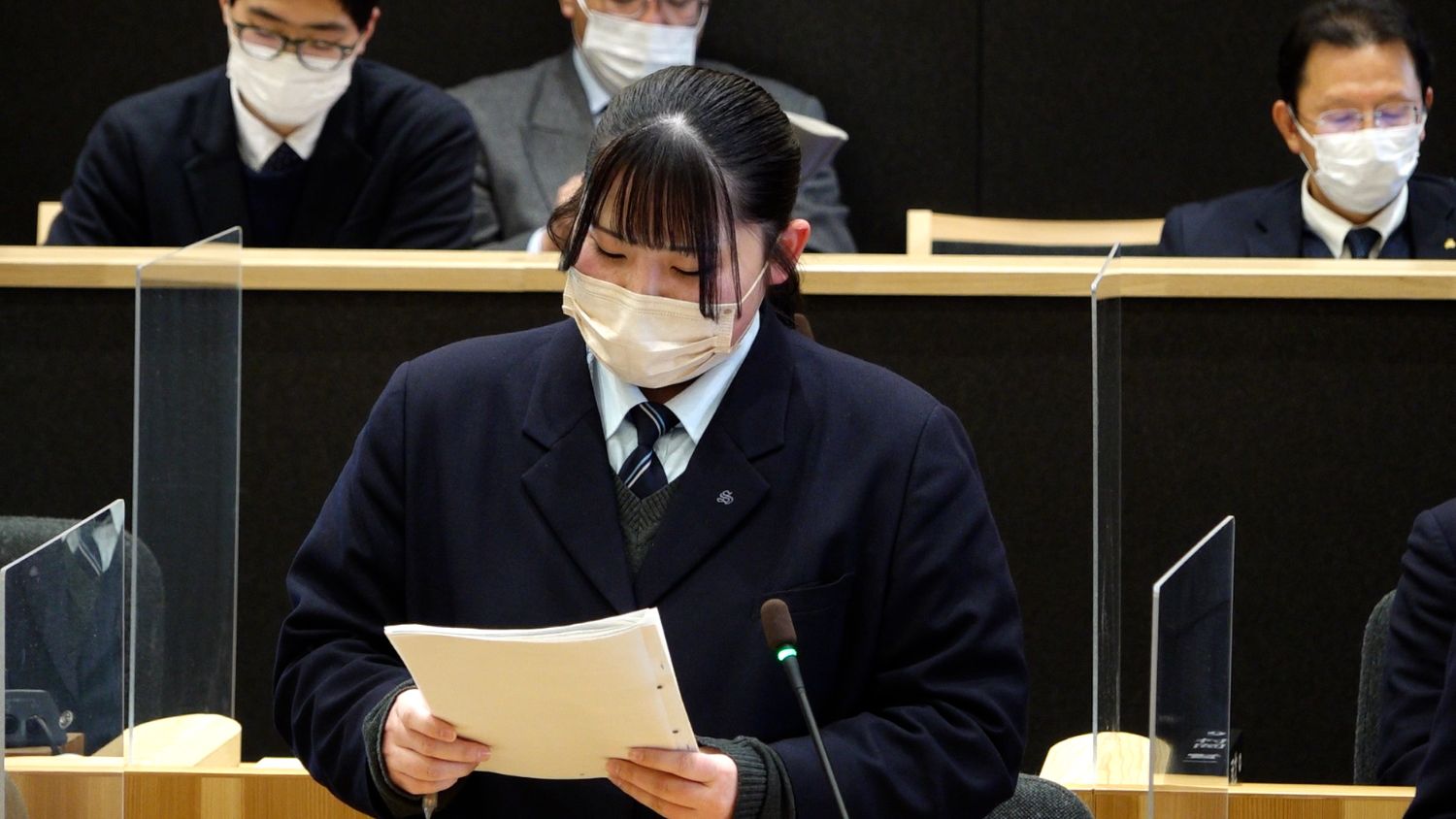 前方に立つ女子学生が発表原稿を読みながら発表しており、後方ではスーツ姿でマスクを着用した複数の人々が資料を見ながらその発表を静かに聞いている様子の写真