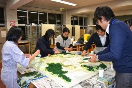 数人の参加者が模型の上に緑色のスポンジ素材をまいて山や森林の再現作業を行っている様子の写真