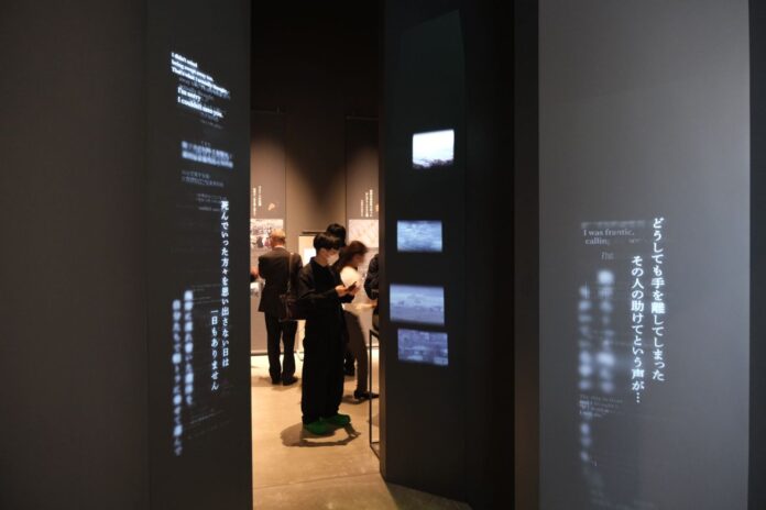 薄暗い展示室の中で、壁に英語と日本語の津波被災者の証言が光で投影されており、来場者が展示を熱心に読んでいる様子が写っている写真