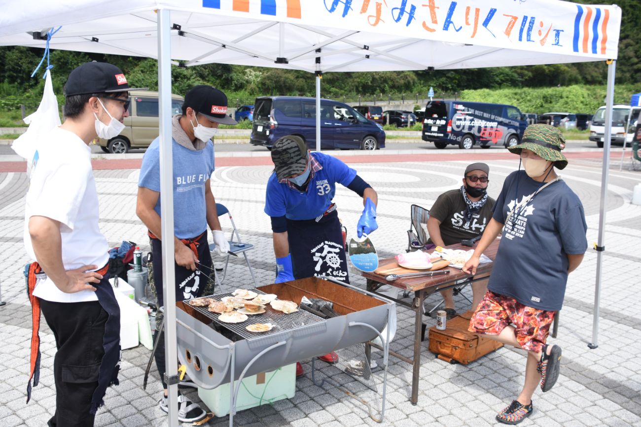 屋外のテント下で、数人が焼き台で貝を焼いて選手たちへ提供している写真