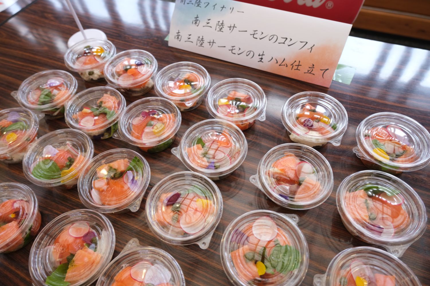 透明な蓋つきのプラスチック容器に盛り付けられたサーモンの刺身や野菜の小鉢が多数テーブルに並んでいる写真