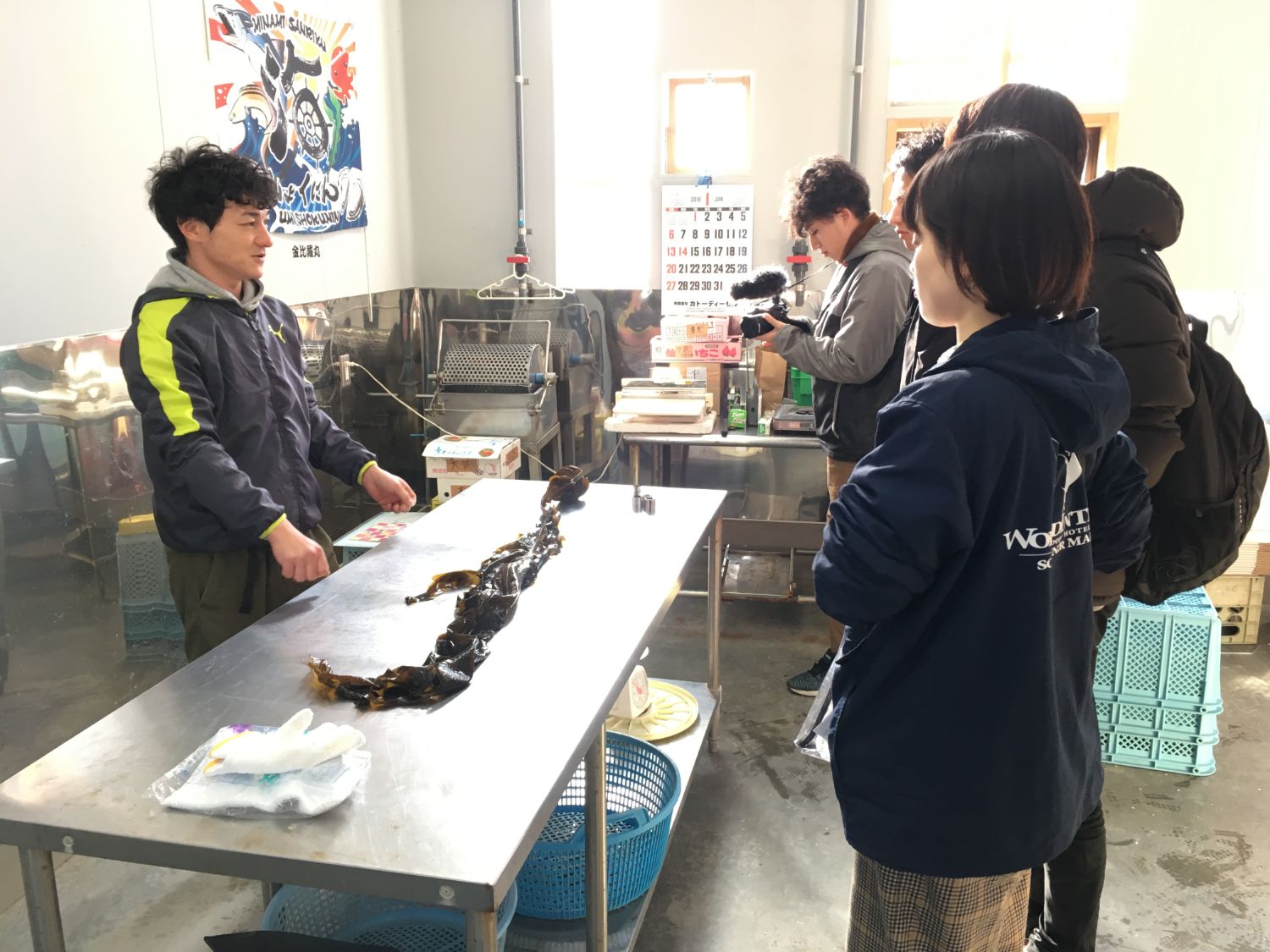 男性がテーブルに広げた昆布を見学者たちに説明しており、周囲には撮影者や工場の機械などがある写真