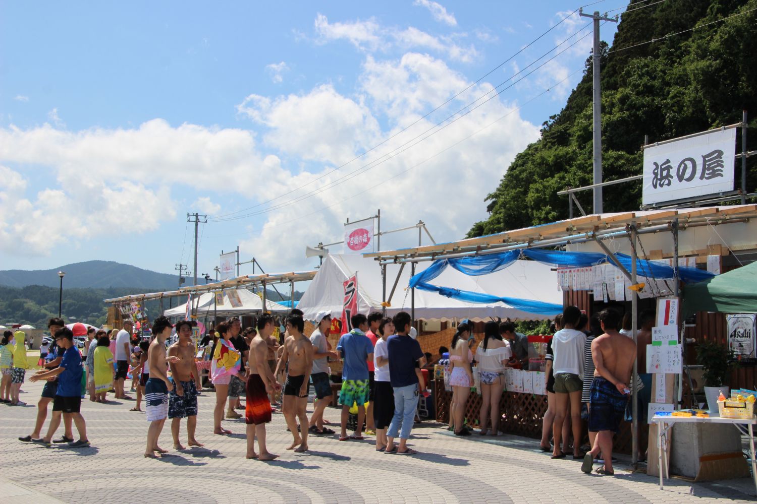 海辺に並ぶ屋台の前で、食べ物や飲み物を求めて多くの人々が列を作っている様子を写した海の家の写真