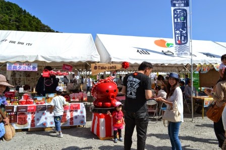 白いテントが並ぶ屋台会場で、「タコcafé」と書かれたブースの前に赤いタコの置物があり、女の子が前に立ち撮影している親子連れが楽しそうに過ごしている写真