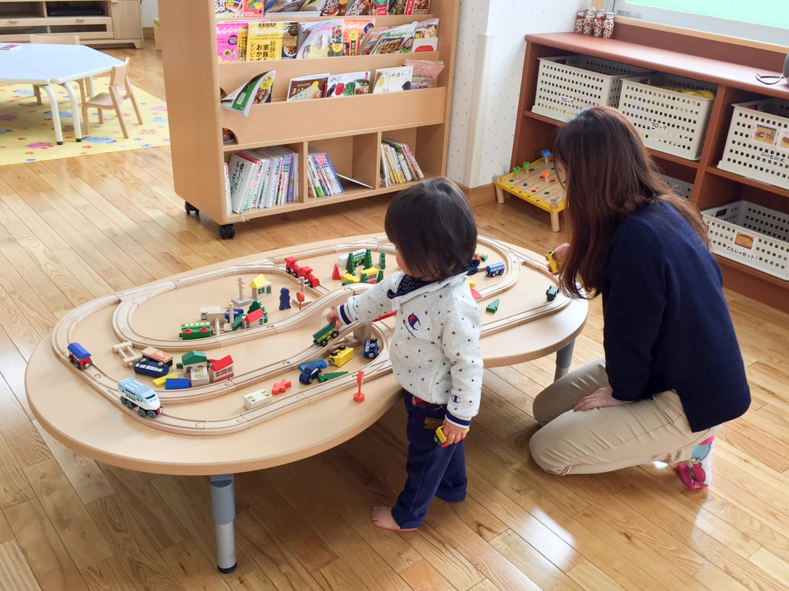 たくさんの本が入った本棚の手前に置かれたテーブルの上で、設置された模型の木のレールに車を置いて遊ぶ菅原さん親子の写真