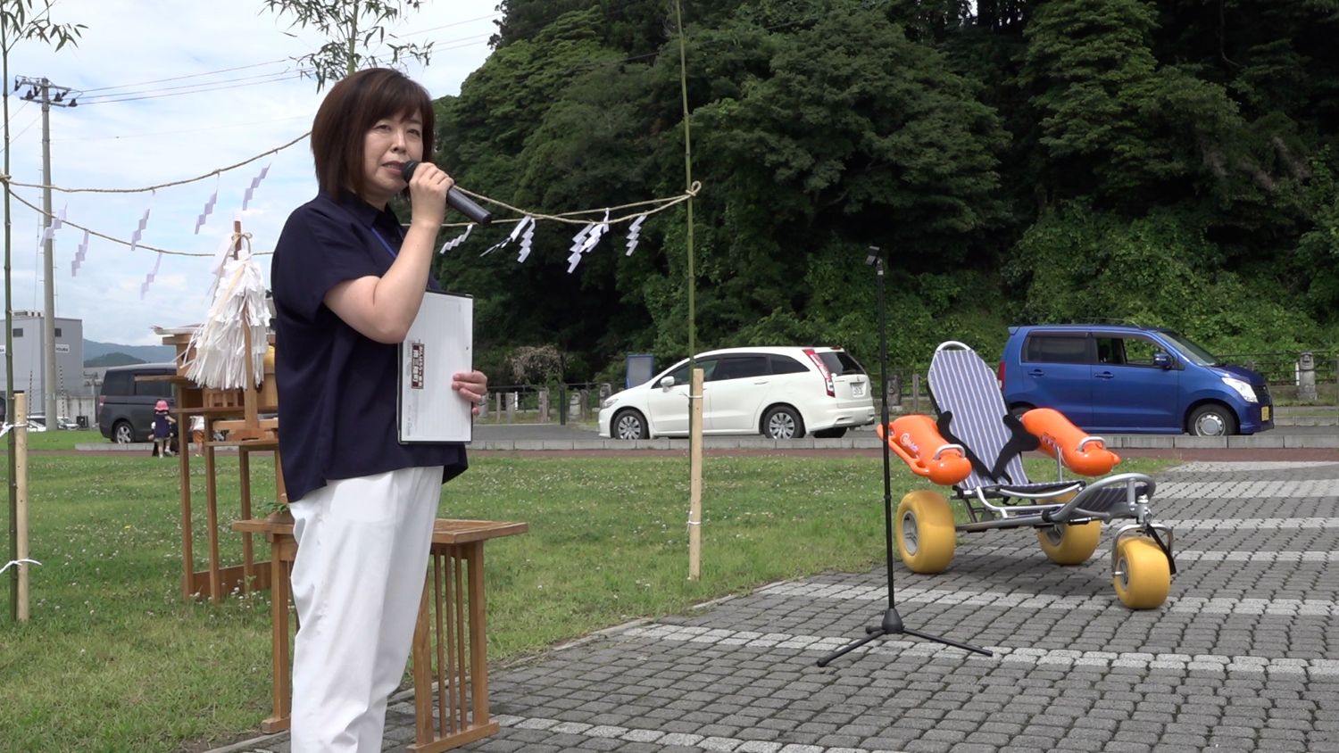 背後に神事の祭壇としめ縄、青とオレンジの車いす型ビーチ用具が見える芝生の広場で女性がマイクを持ってスピーチをしている写真