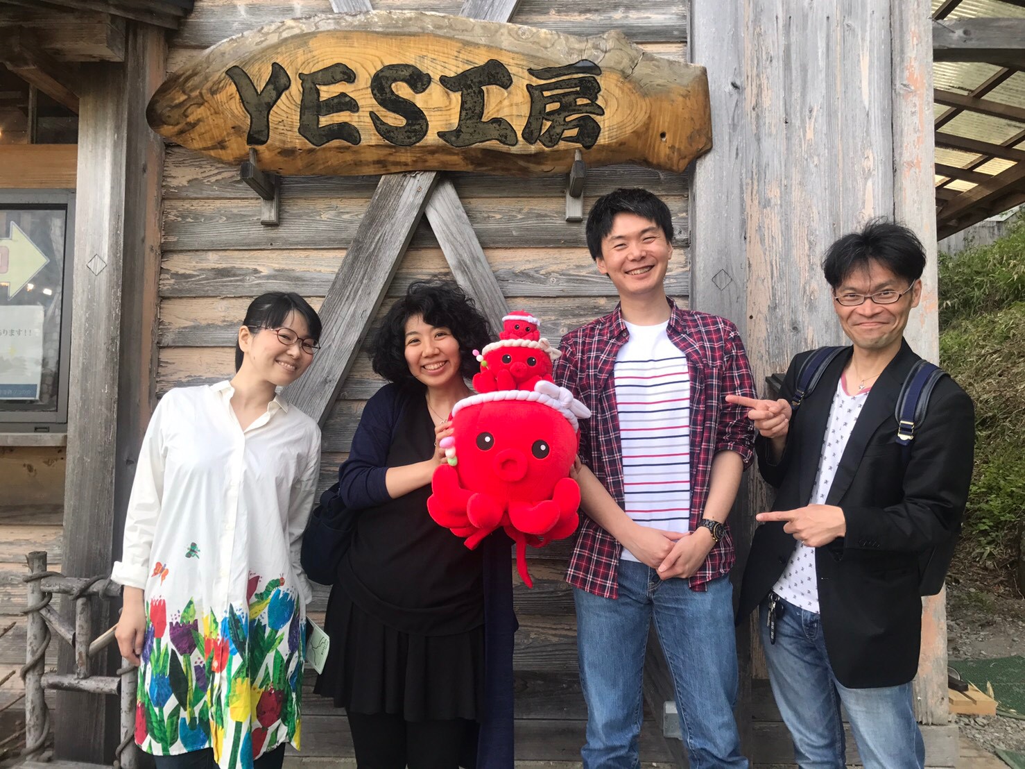 「YES工房」の看板の前で、女性2人と男性2人が横一列に並び、左から2番目の女性がオクトパス君のぬいぐるみを持って笑顔で並んでいる写真