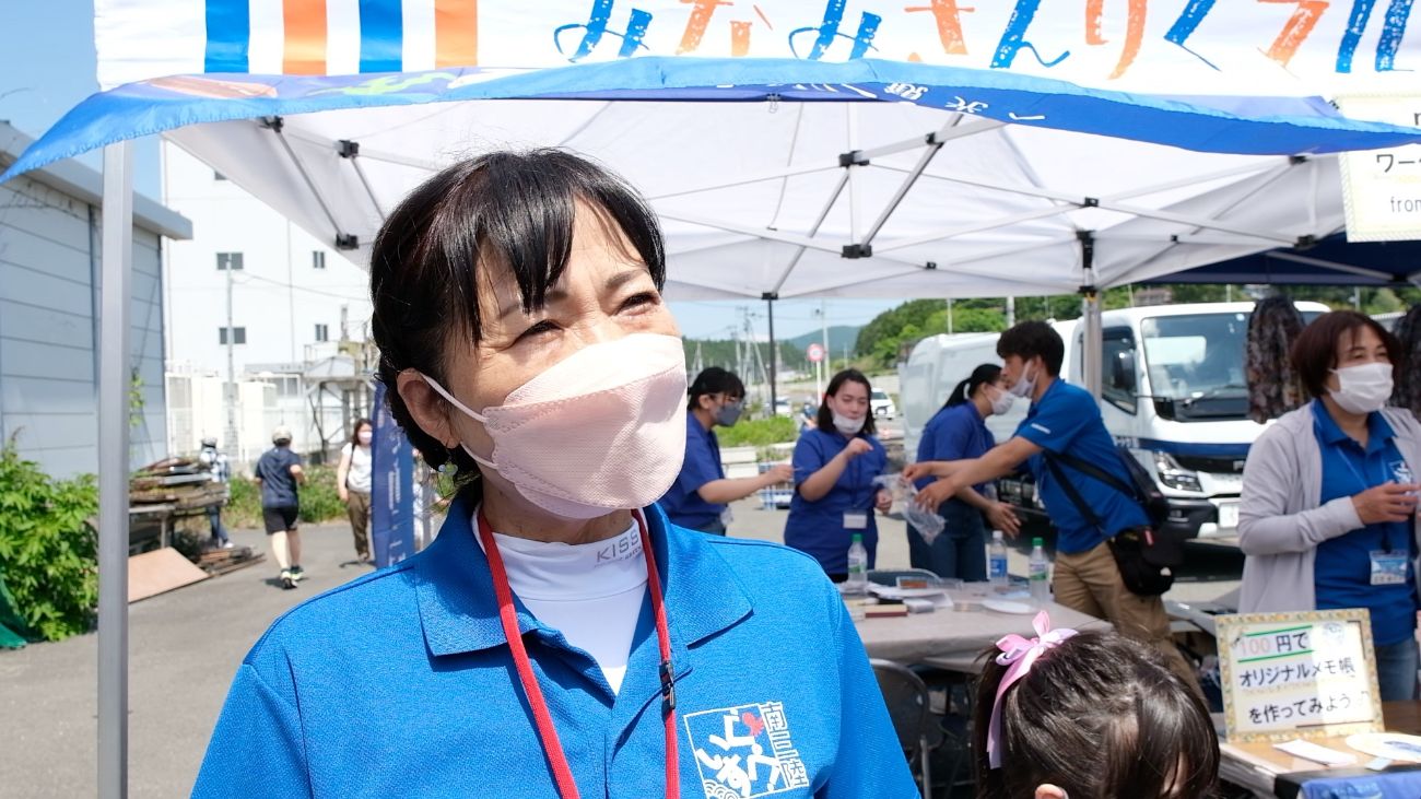 復興市の屋外ブースで青いユニフォームを着た上一枝さんが、笑顔でインタビューに答えている写真