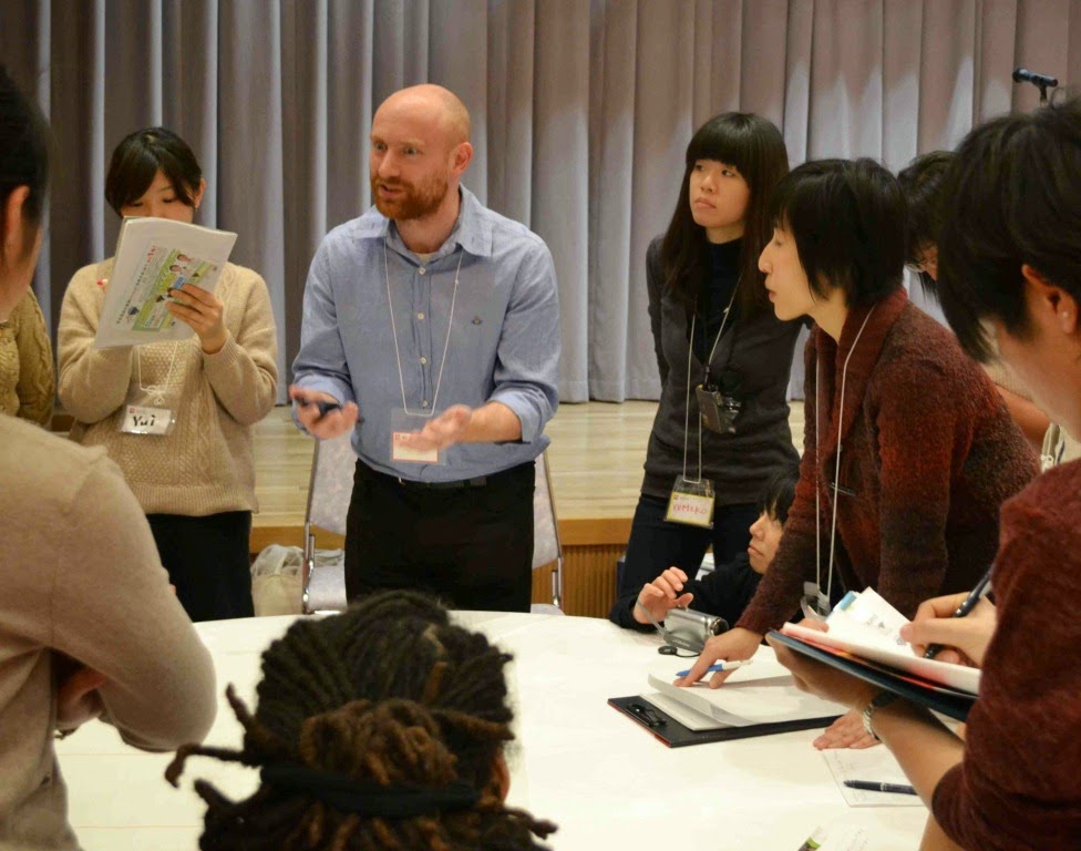 外国の男性が資料を手に持ちながら、円形のテーブルの周りに並んでいる参加者の人たちに説明をしている様子を写した写真
