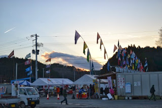 夕暮れ時の商店街に色とりどりの旗がはためき、屋台で人が作業している様子が見える写真