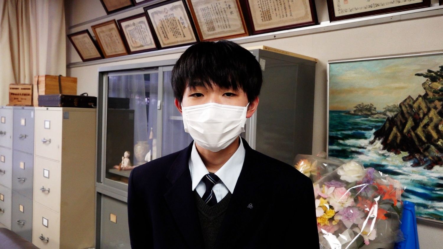 ガラスケースや花束、美術作品の絵が並ぶ部屋で、マスクをつけた制服姿の男子高校生の写真