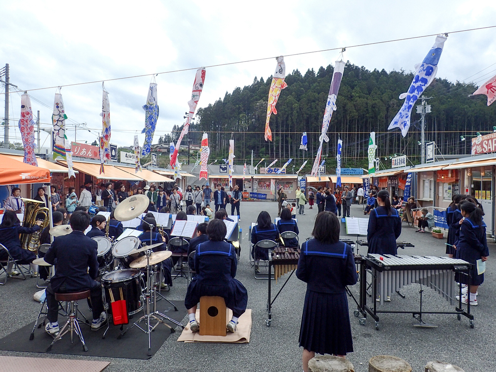 ロープに並んだたくさんのこいのぼりがたなびく南三陸さんさん商店街で、吹奏楽部の演奏を観客が囲むように立ち見している写真