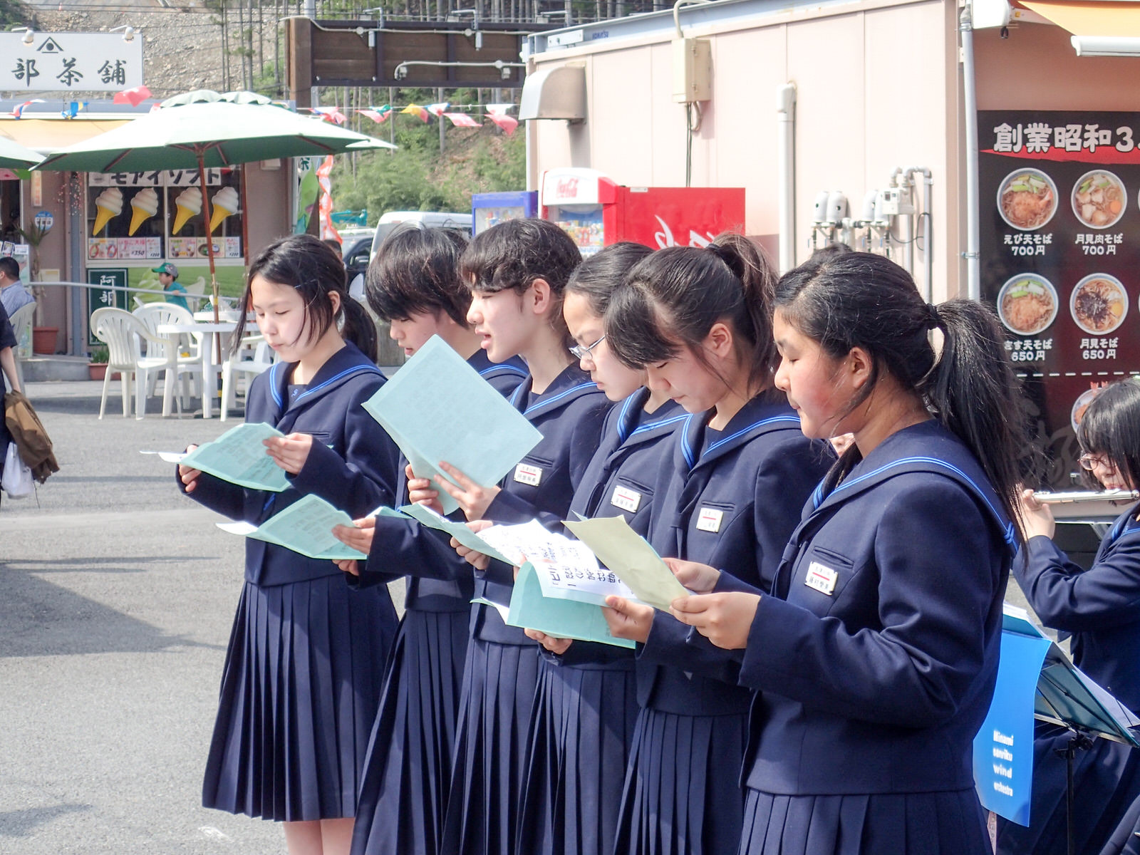 制服を着た女子中学生たちが歌詞カードを持って合唱している様子の写真