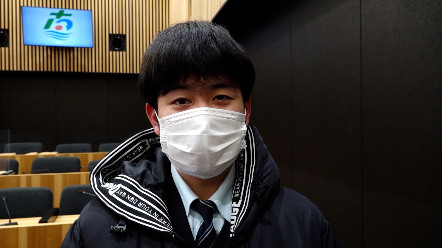 議場内で、黒い防寒着を着てマスクを着けた男子高校生がカメラの前に立っている写真