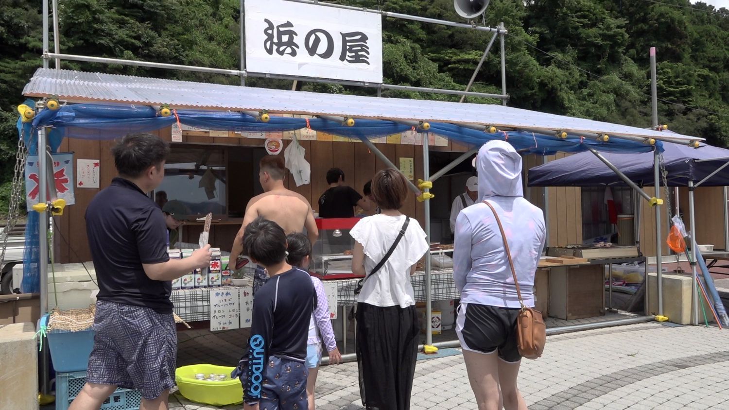 木製の建物に透明な波板屋根と青いネットをかけた仮設風の売店「浜の屋」に、水着姿や夏服の家族連れなど数人が並んでいる写真