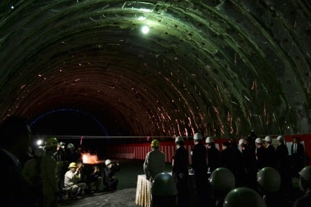 ヘルメットを着用した人々が一か所だけ灯りが灯された暗いトンネル内に集まっており、代表者が横一列に整列して立ち、左側に座っている人たちがカメラを構え、少し離れた場所の地面から花火の様な炎が現れている貫通式の様子の写真