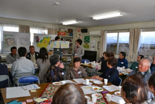 壁に貼られた地図や資料をもとに、前に立つ話し手が説明を行い、それを囲む参加者たちが真剣に聞き入っているお茶っこ会の様子の写真