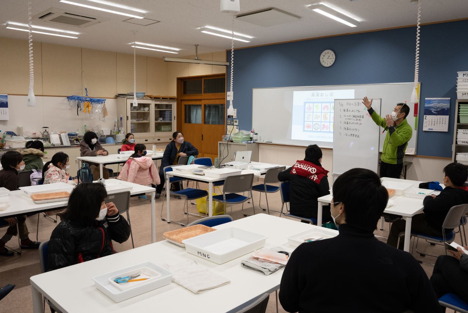 関係者の男性がホワイトボードとスライドを使って授業を行い、子どもたちが座って説明を真剣に聞いている写真